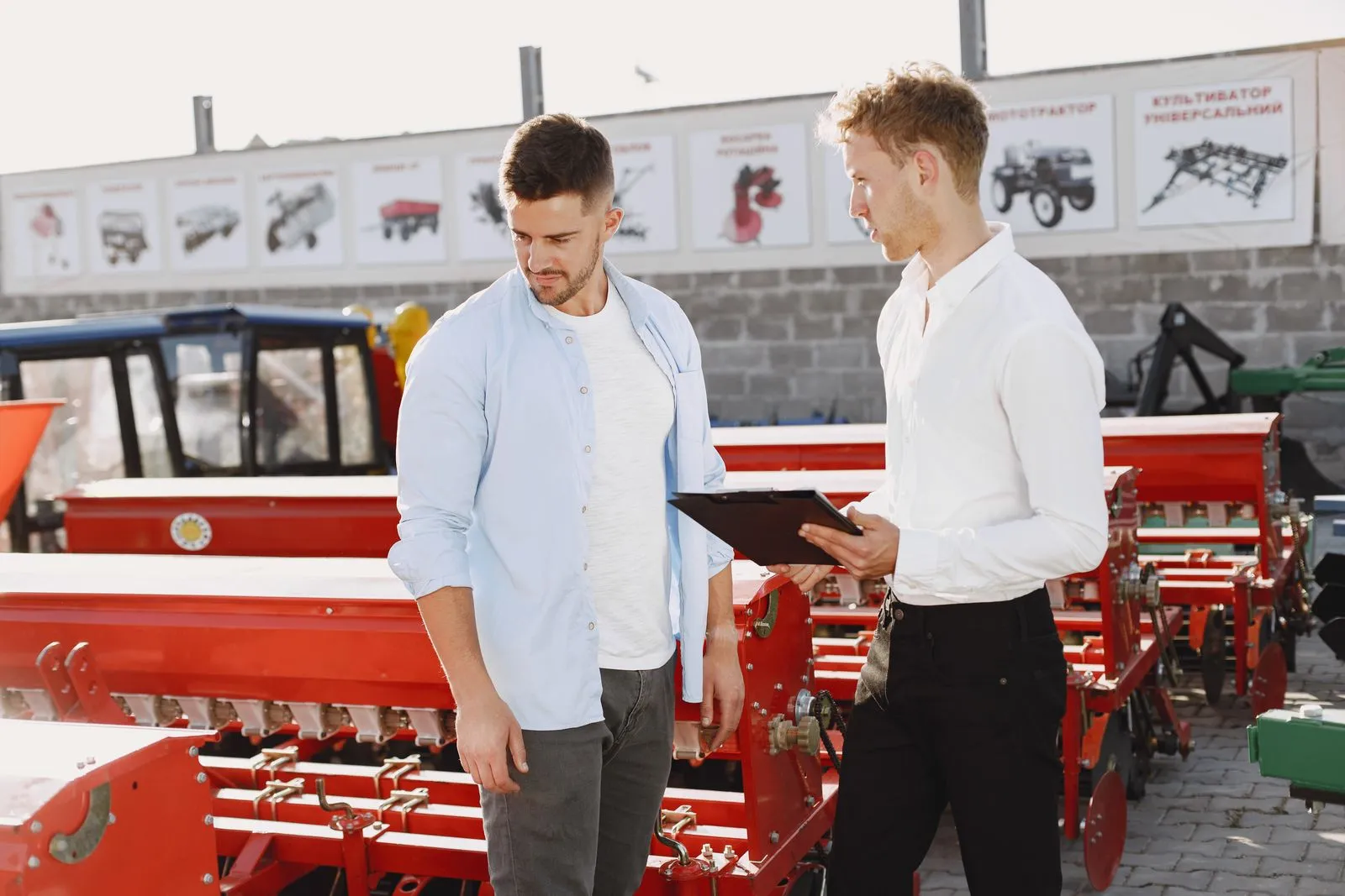 A buyer and a sales rep with a clipboard reviewing paperwork beside new farm machinery at an equipment dealer